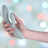 A hand holding the Svakom Chika Rabbit Vibrator, an app-controlled turquoise grey device crafted with liquid silicone and featuring side buttons. The softly blurred background with pastel-colored circles of light enhances the sleek design.
